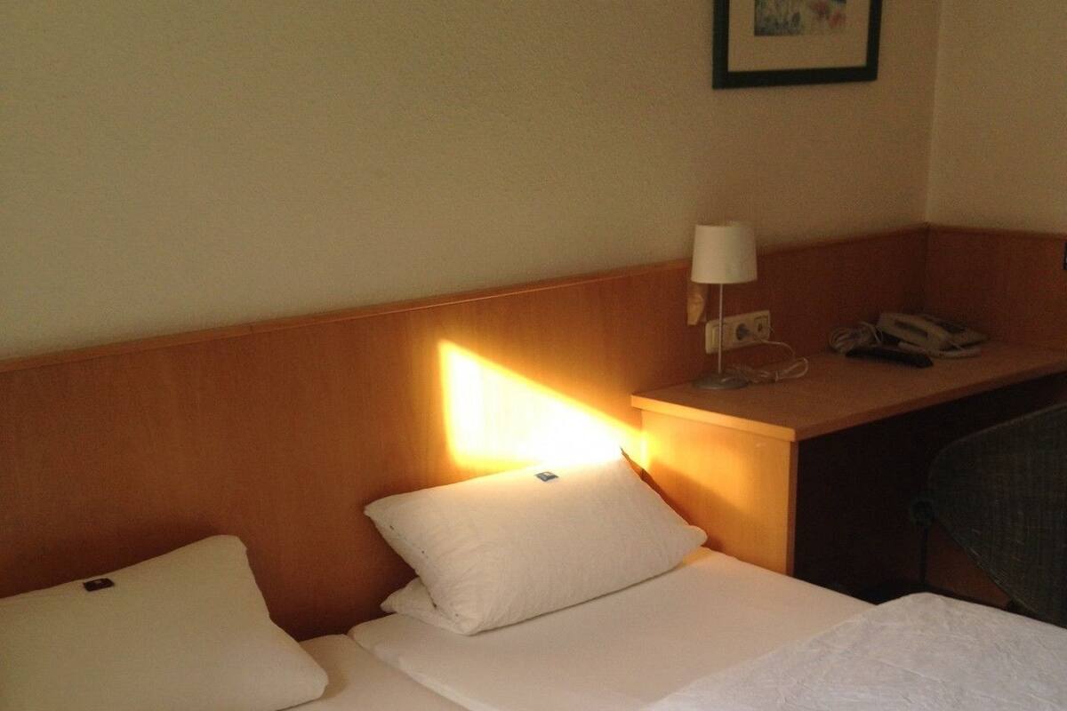 A hotel room with a bed, pillows, a bedside lamp, a desk with a phone, and a framed picture on the wall.