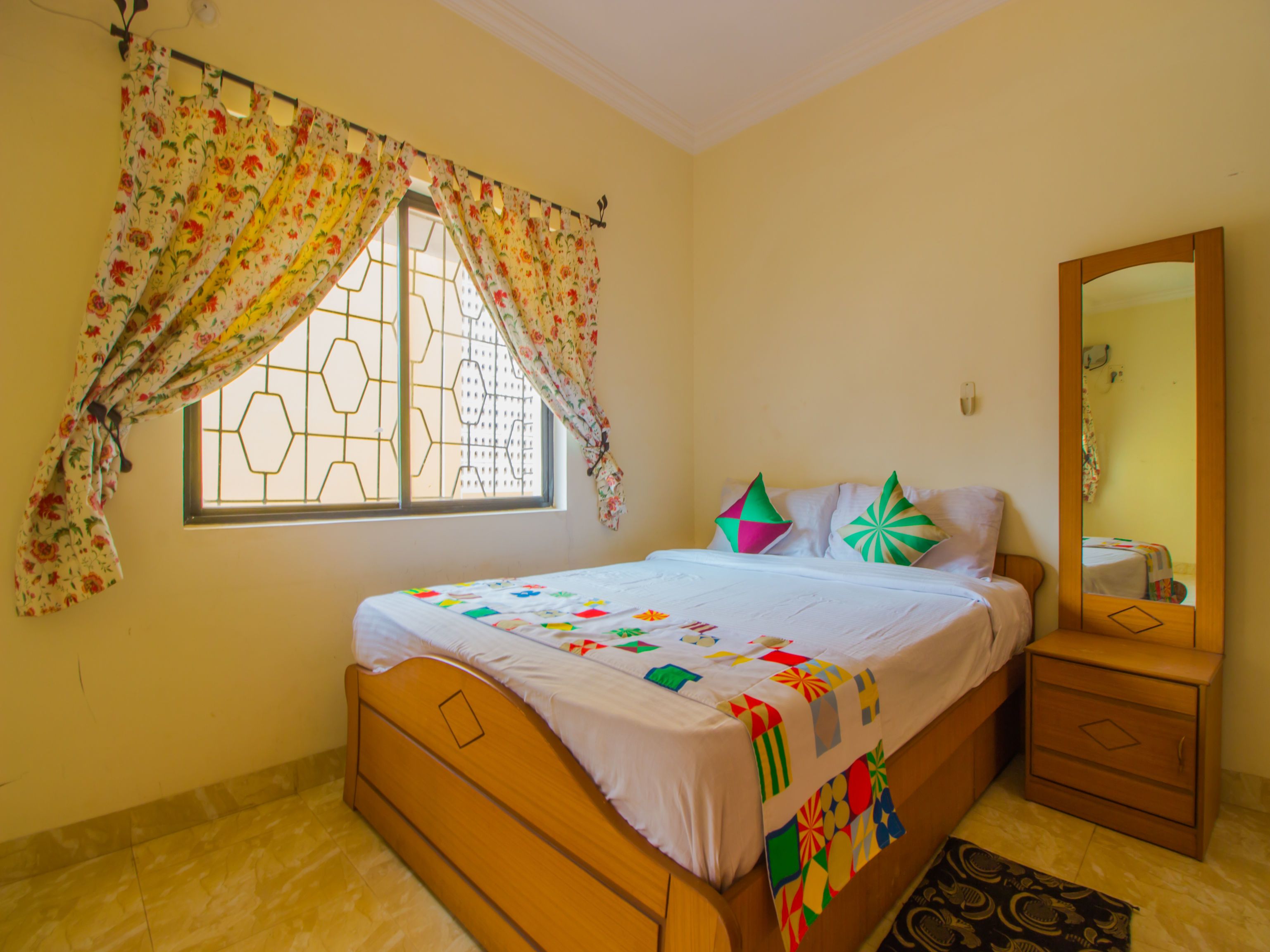 A bedroom with a bed, a nightstand, a mirror, and a window with floral curtains.