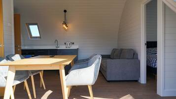 A compact, well-lit room with a dining table, chairs, a sofa, and a kitchenette.