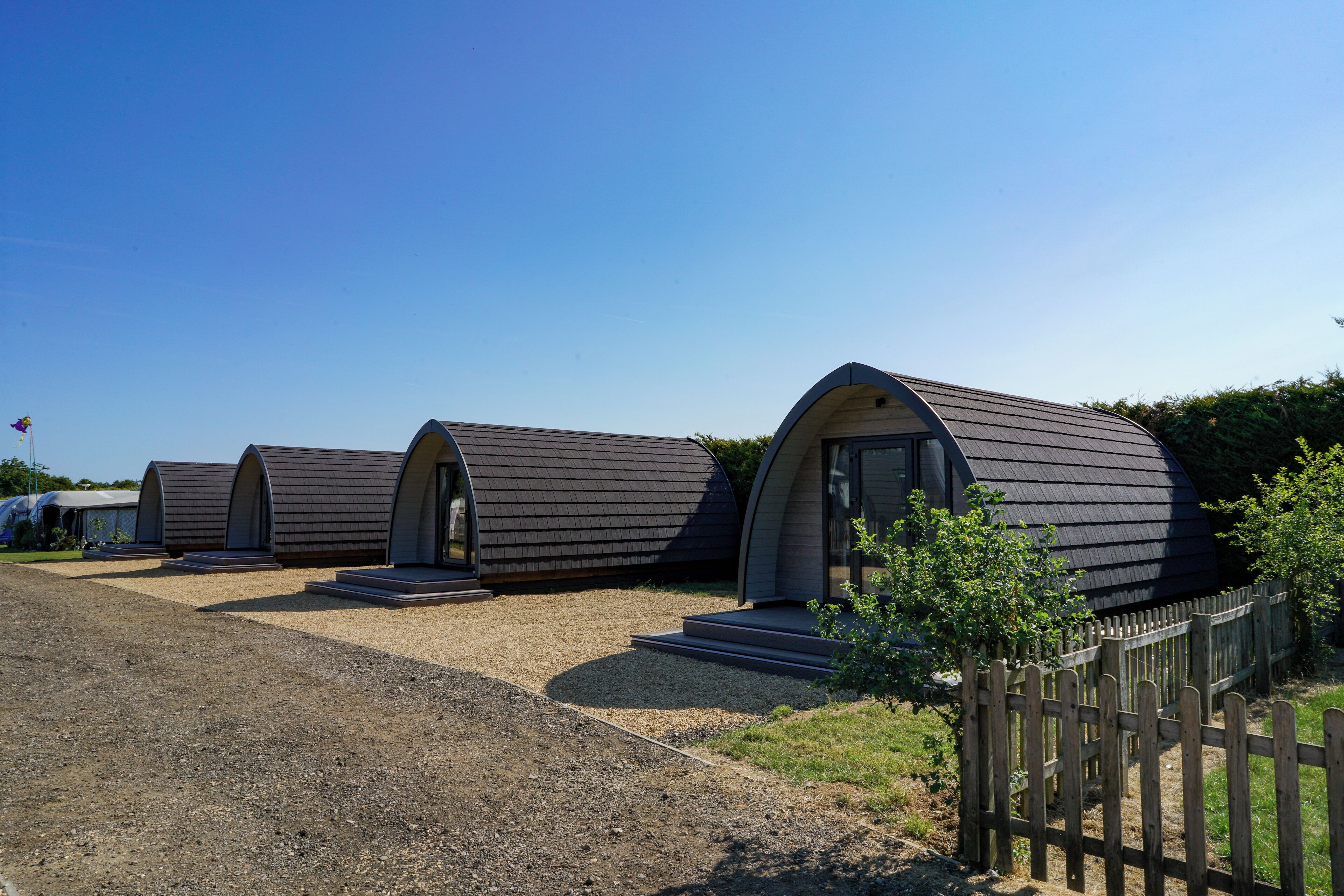 A row of modern, curved-roofed cabins with a gravel driveway and a wooden fence.