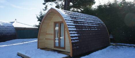 Uma cabana pequena de madeira, em formato semicircular, com porta e janelas de vidro, rodeada por neve.