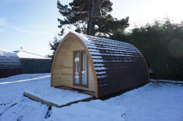 A small, wooden, semi-circular cabin with a glass door and windows, surrounded by snow.