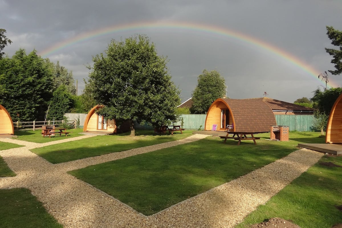 Une série de cabanes en bois en forme de dôme, dotées de tables de pique-nique, disposées dans un espace verdoyant, avec un arc-en-ciel visible en arrière-plan.