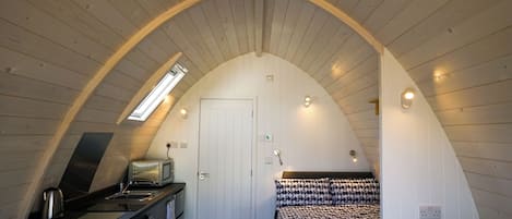 A cozy attic bedroom with a kitchenette, a bed with patterned bedding, and a sloped ceiling with wooden beams.