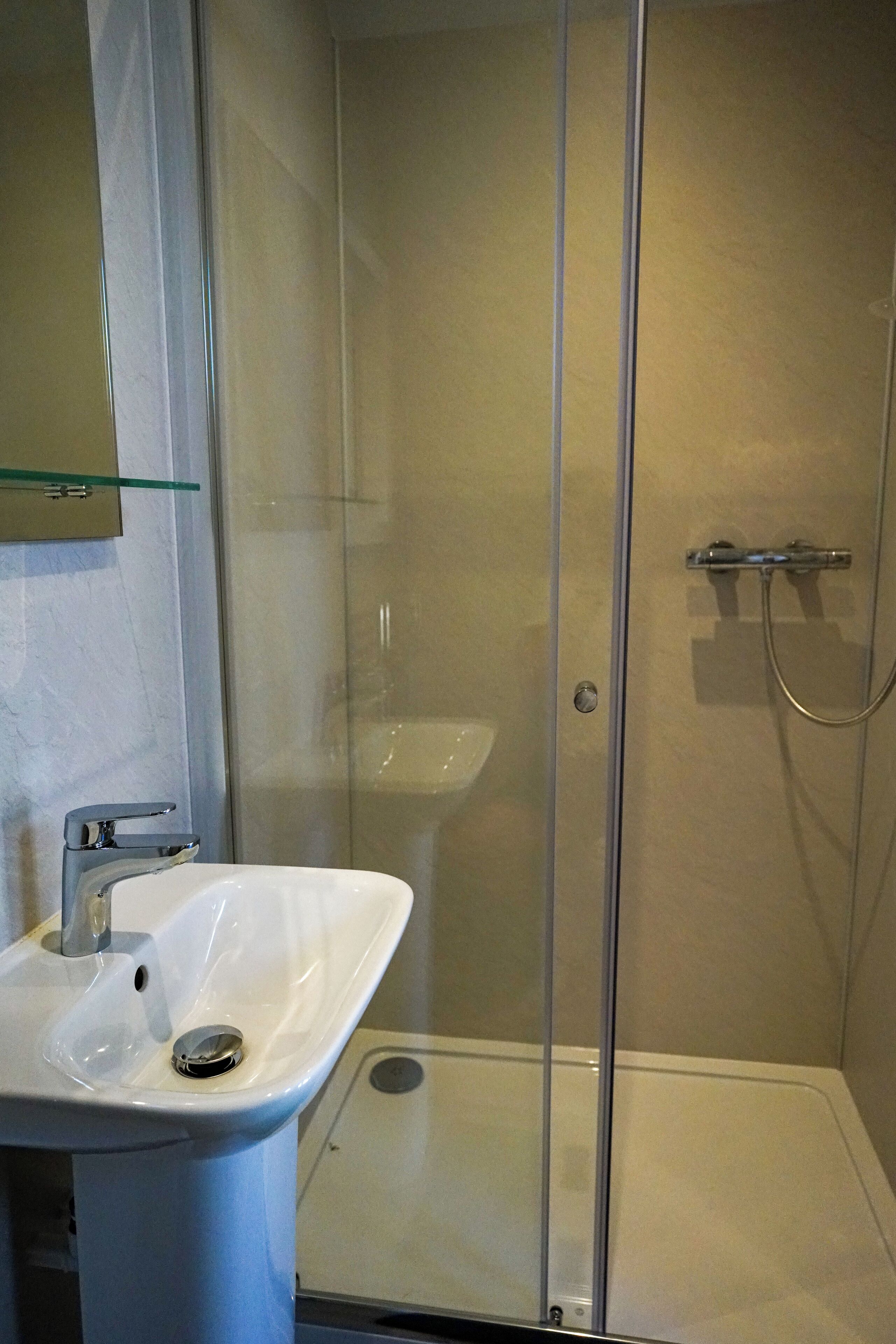 A bathroom with a glass-enclosed shower, a white sink, and a glass shelf above the sink.