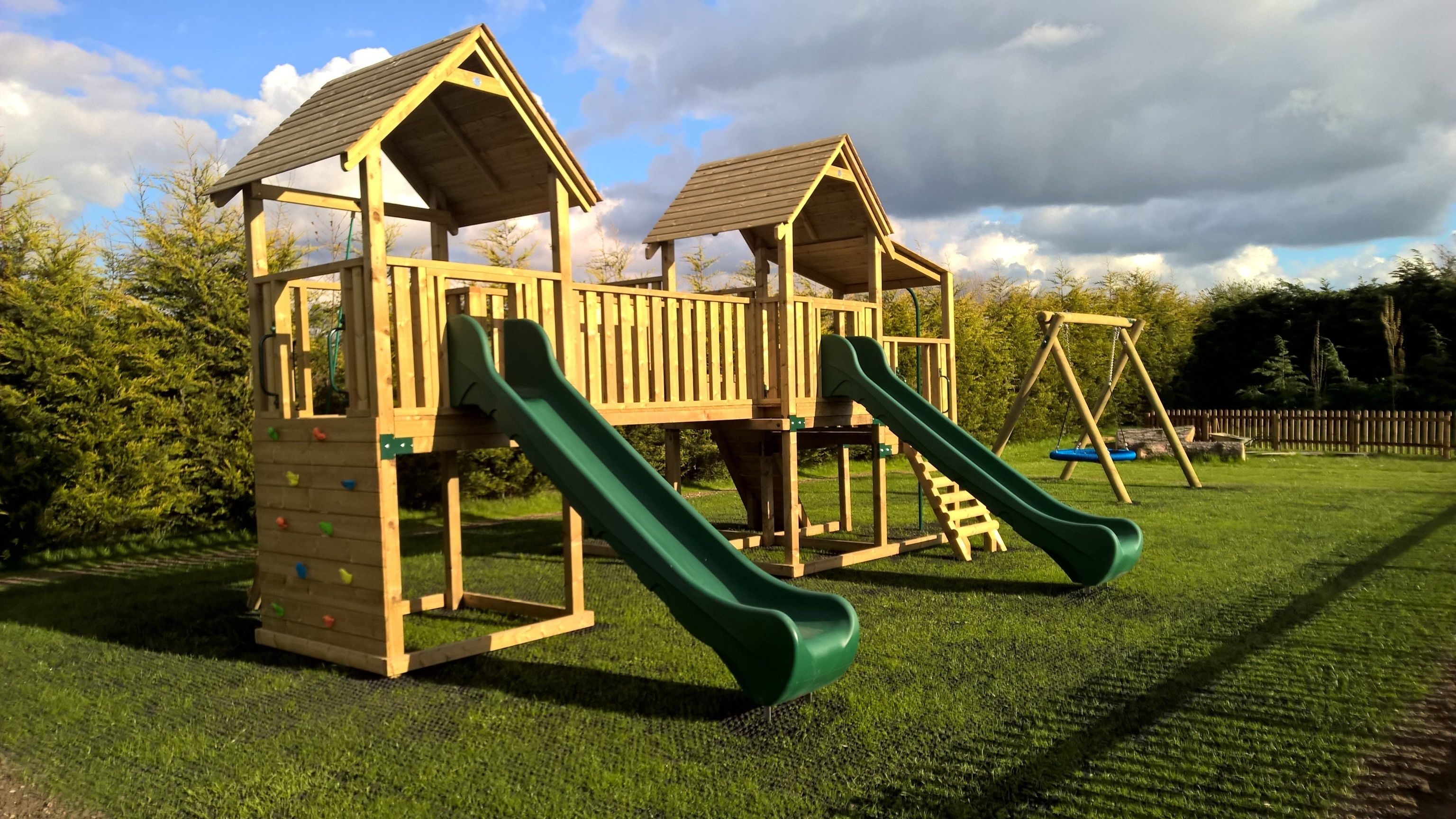 A wooden play structure with two slides and a climbing wall, set on a grassy field with trees in the background.