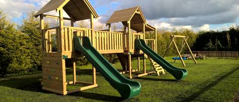 A wooden play structure with two slides and a climbing wall, set on a grassy field with trees in the background.