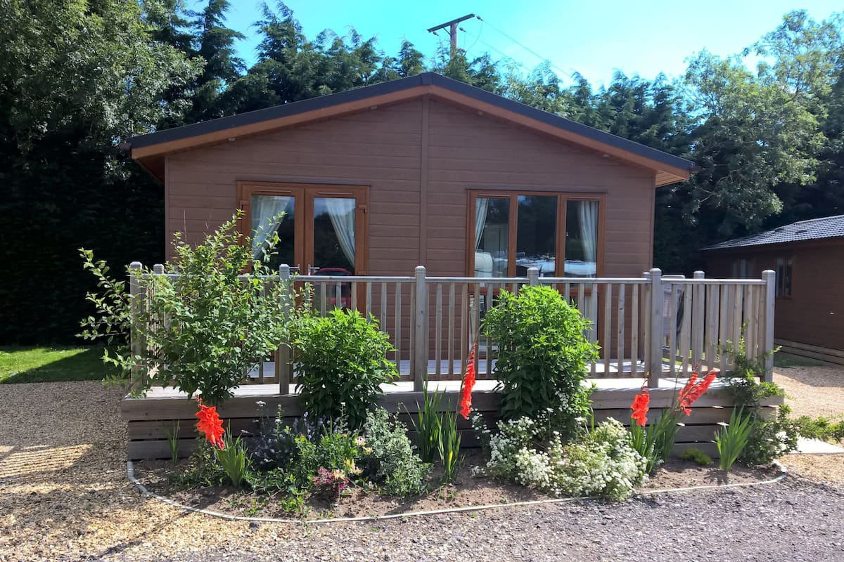 Une petite cabane en bois d’un seul étage, dotée d’une terrasse, entourée d’une allée de gravier et de diverses plantes.