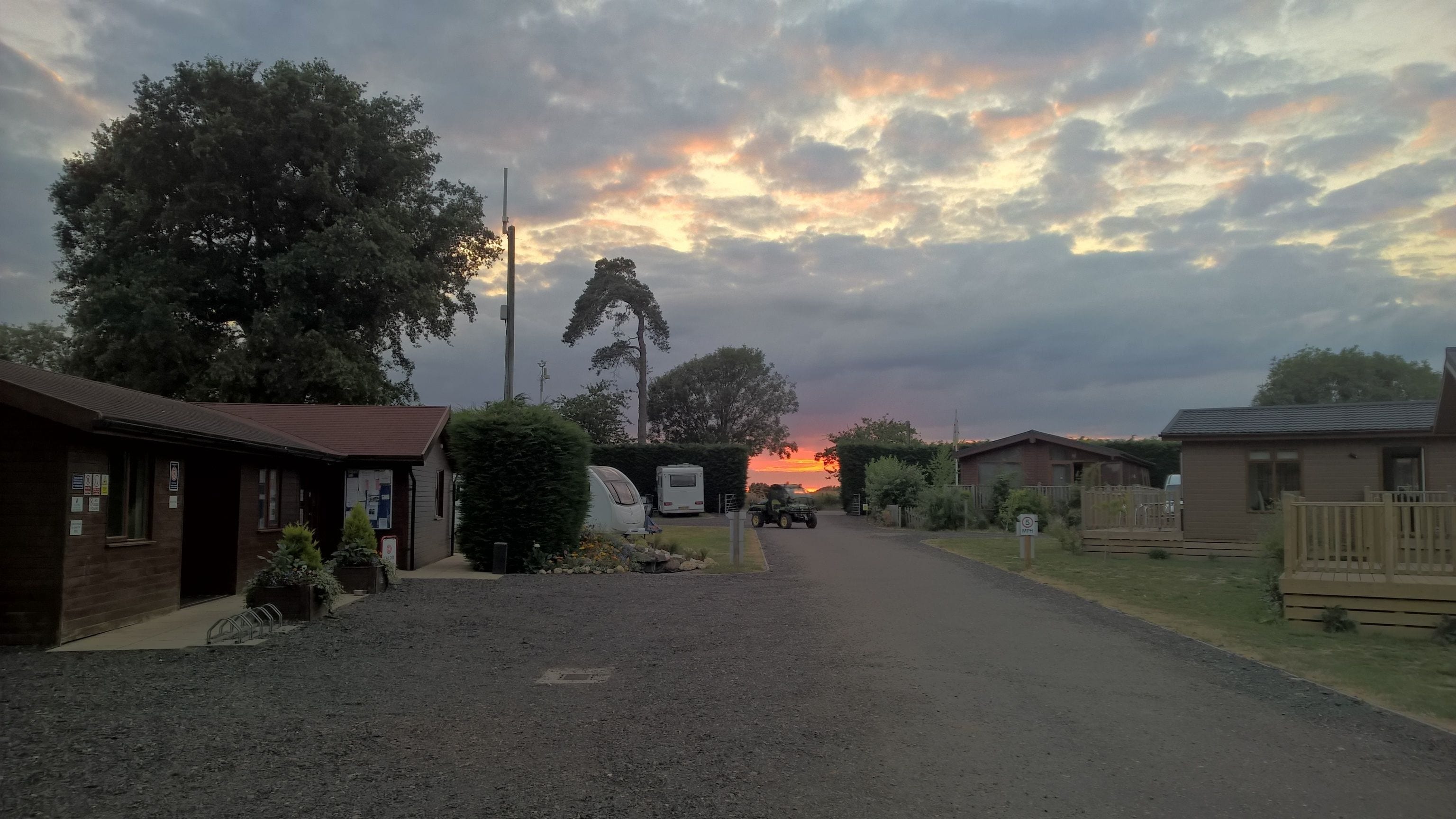 Un camino rural con un camino de grava que conduce a una hilera de edificios, incluyendo una pequeña construcción de ladrillo y una casa de madera.