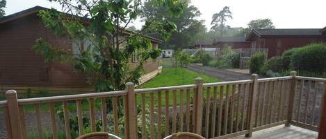 A wooden deck with a round table and wicker chairs, overlooking a garden with a wooden building and a pathway.