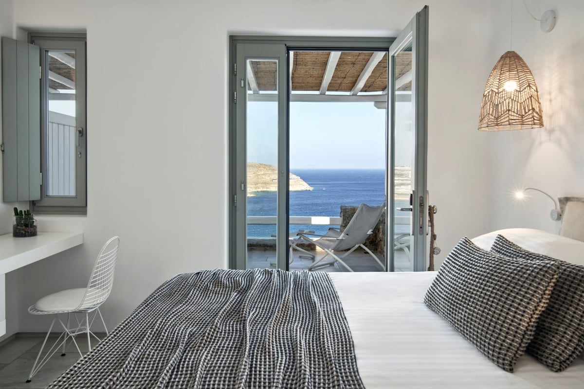 A bedroom with a bed, a chair, a bedside table, a lamp, and a view of the sea.