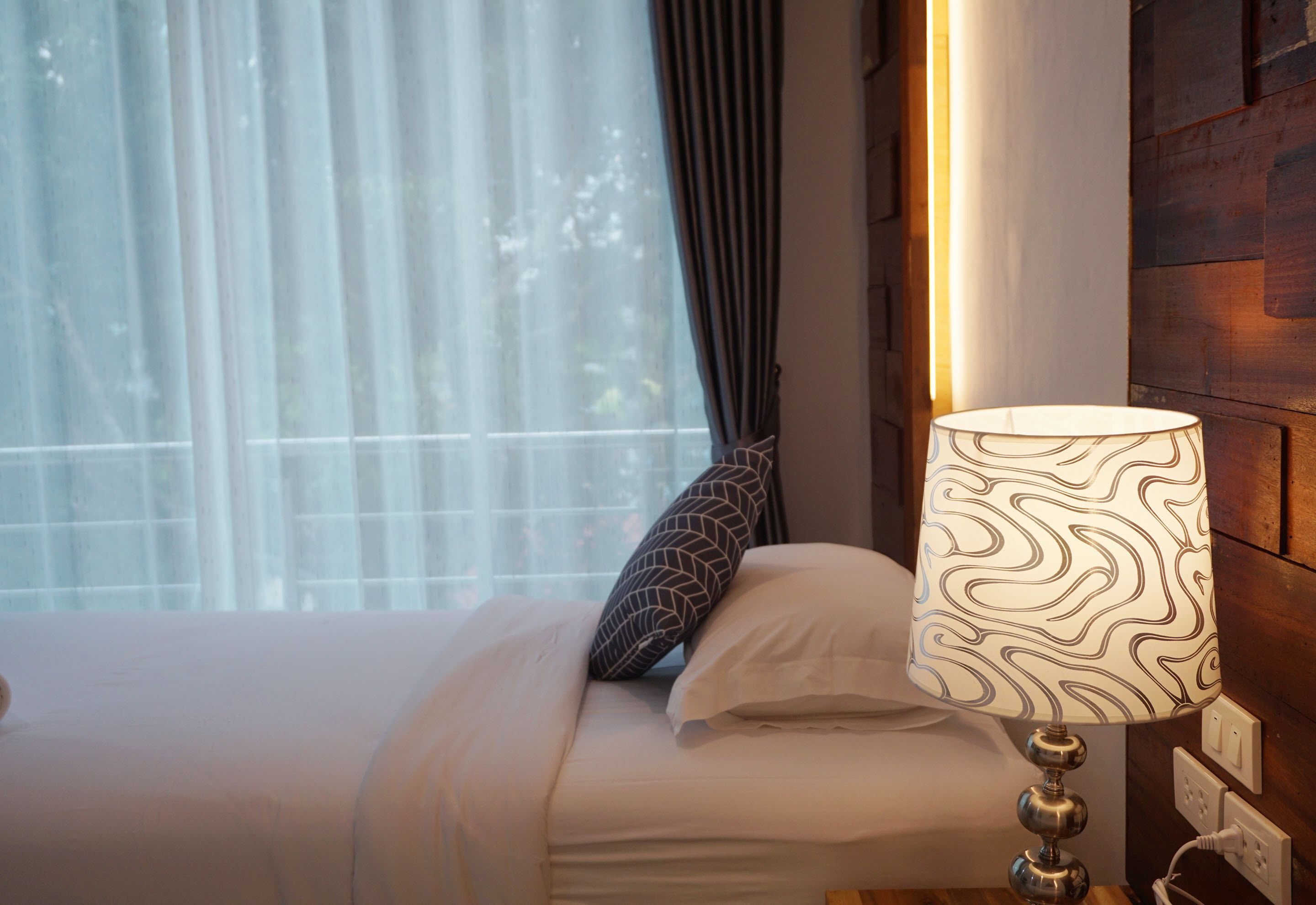 Uma cama bem arrumada com uma luminária estampada, uma janela com cortinas finas e uma parede de madeira.