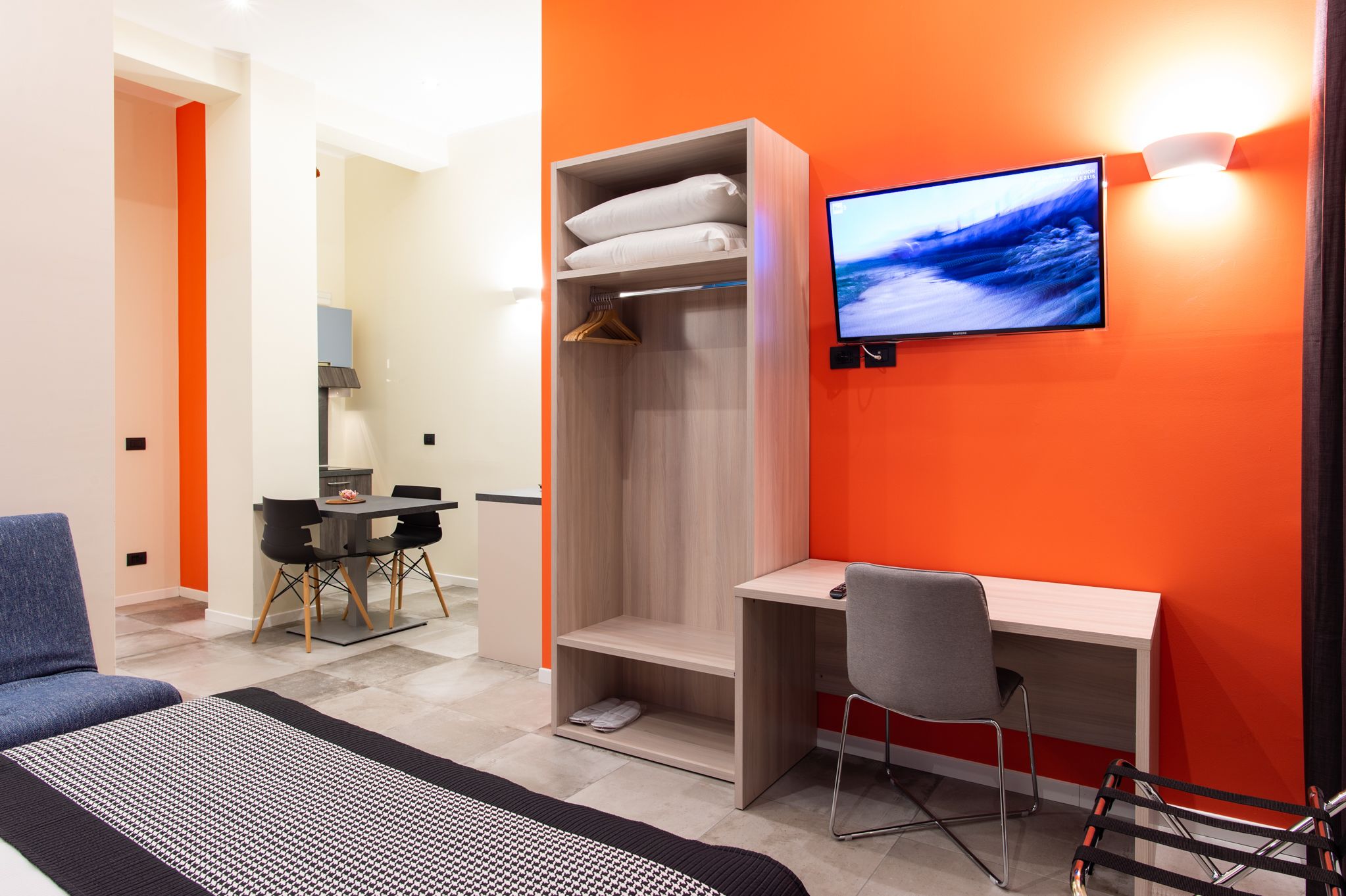 A modern hotel room with a wooden desk, a grey chair, a wardrobe, and a TV mounted on the orange wall.