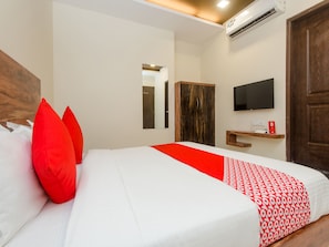 A neatly made bed with white and red bedding, a wooden wardrobe, a mounted TV, and a small shelf with a red object on it.