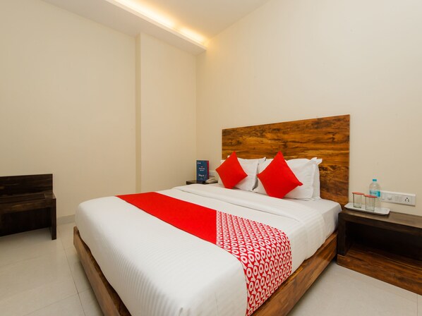 A neatly made bed with a red and white bedspread, a wooden headboard, and a bedside table with a bottle and glasses. - OYO 11747 Hotel Sai Comforts (Vasai)