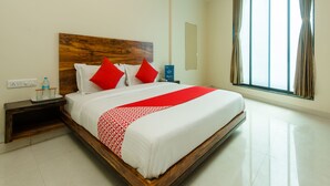 Quarto de hotel com uma cama grande, cabeceira de madeira, mesa de cabeceira com garrafa e copos, espelho e uma janela ampla com cortinas.