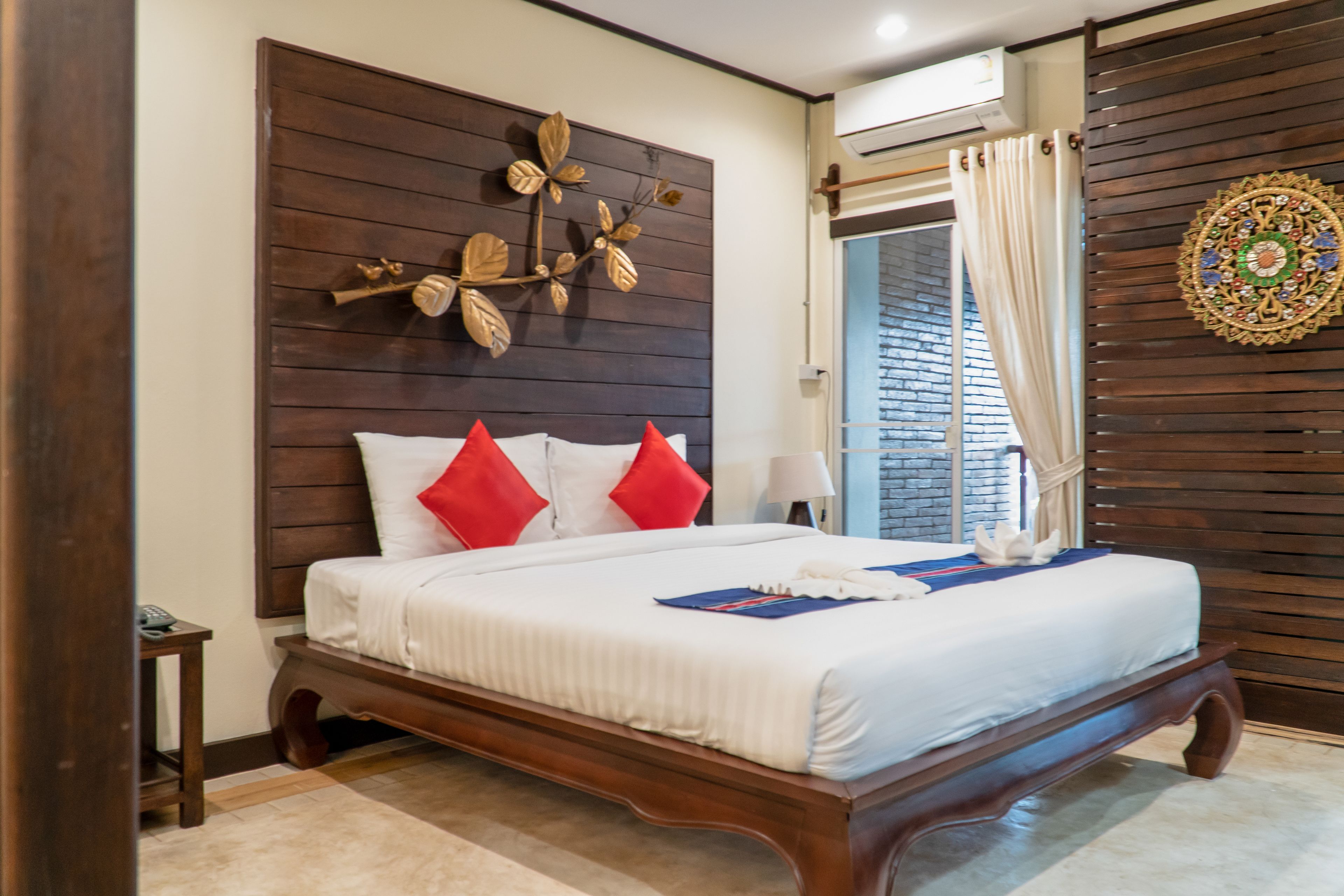 A neatly made bed with white bedding and red pillows, a wooden headboard with a decorative leaf design, and a wall-mounted circular ornament.