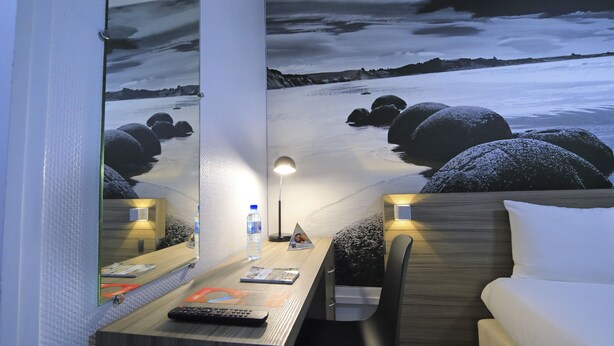 Habitación de hotel con cama, escritorio con lámpara, espejo y una pared decorada con una obra de arte.