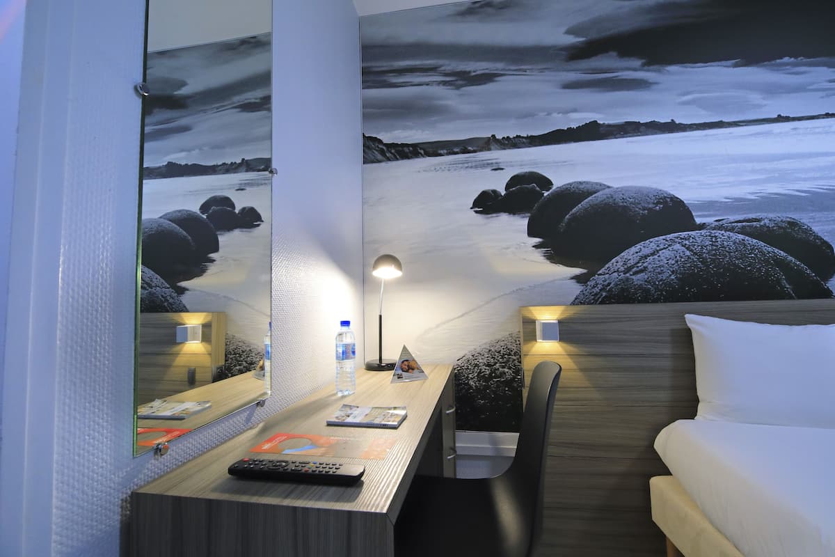 Une chambre d’hôtel avec un lit, un bureau doté d’une lampe, un miroir et un mur orné d’une œuvre d’art représentant un paysage.