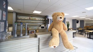 Un oso de peluche grande sobre una barra en una cafetería moderna, con estantes de té y botellas al fondo.