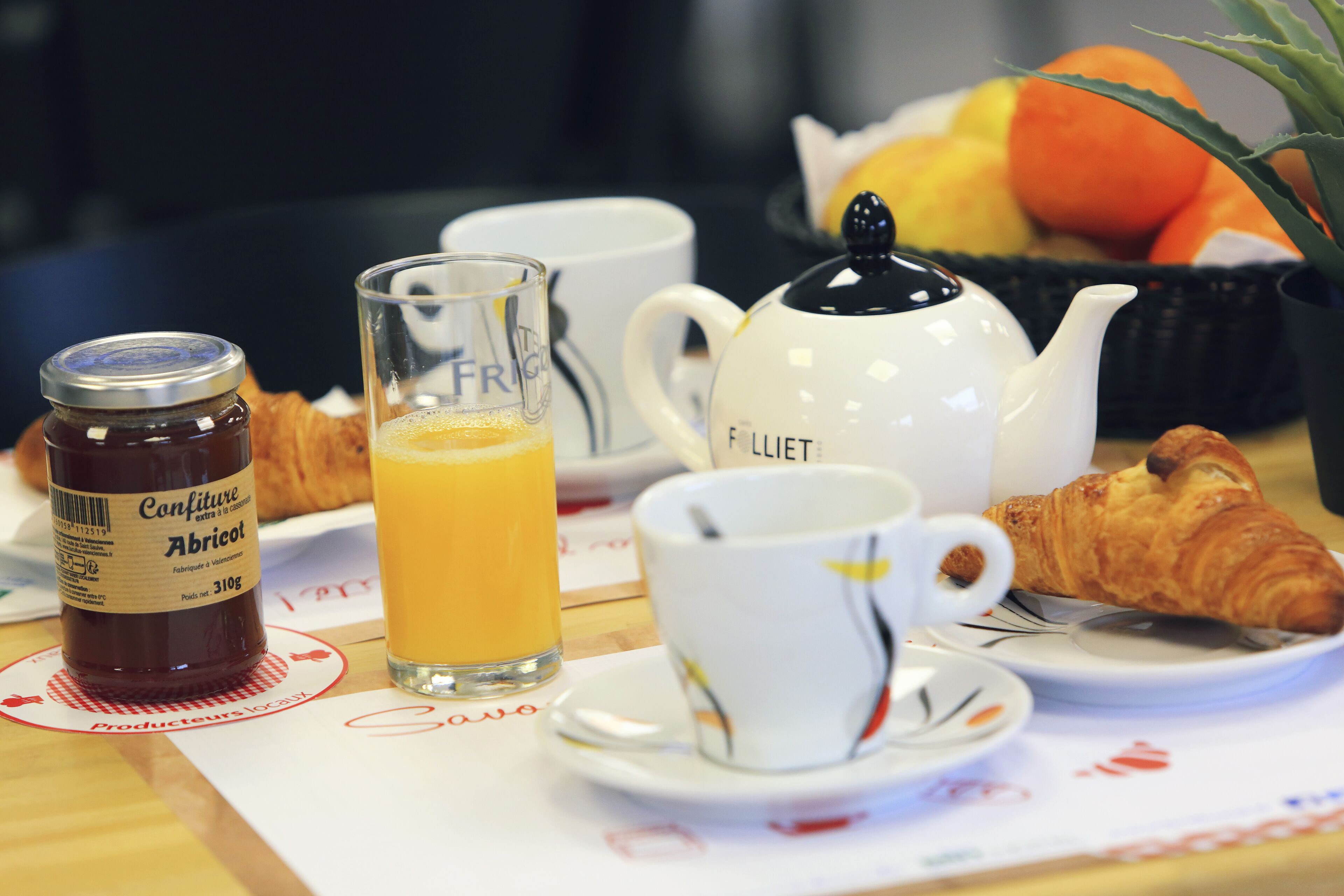 Une table de petit-déjeuner avec un croissant, un verre de jus d’orange, une théière, un pot de confiture et une tasse.