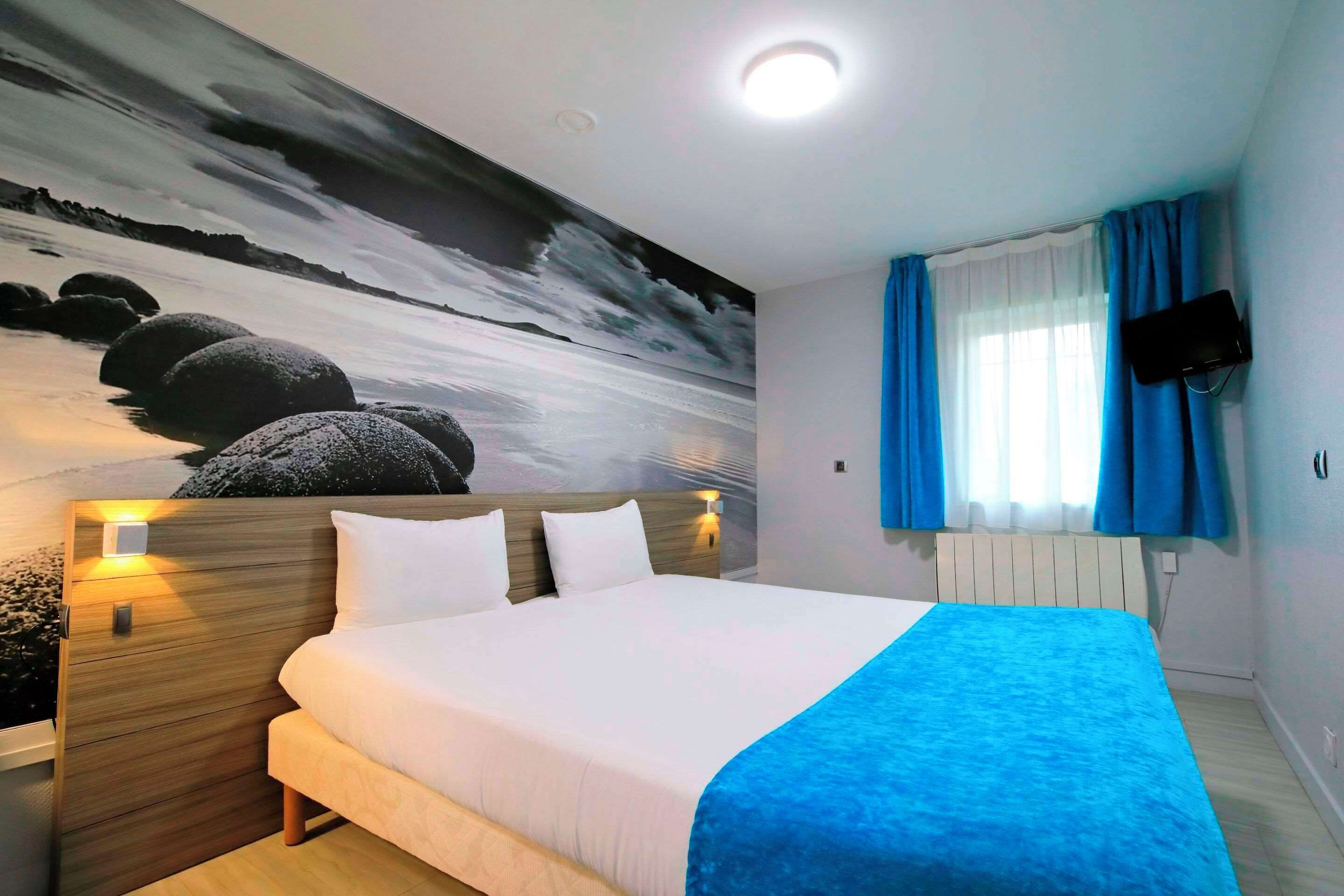 Una habitación de hotel con una cama, una mesita de noche con lámpara, un televisor en la pared y un ventanal con cortinas.