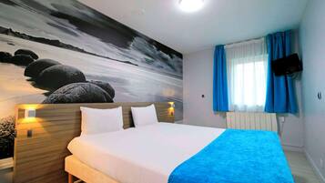 Una habitación de hotel con una cama, una mesita de noche con lámpara, un televisor en la pared y un ventanal con cortinas.