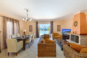 A living room with a dining area, a fireplace, a television, and a view of the pool. - Villa Sofia (Corfu)