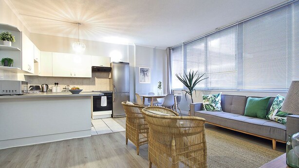 Eine moderne Küche mit weißen Schränken, ein Essbereich mit Korbstühlen und ein Wohnzimmer mit Sofa und großen Fenstern.
