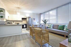 A modern kitchen with white cabinetry, a dining area with wicker chairs, and a living room with a sofa and large windows. - Perspectives (Cape Town)