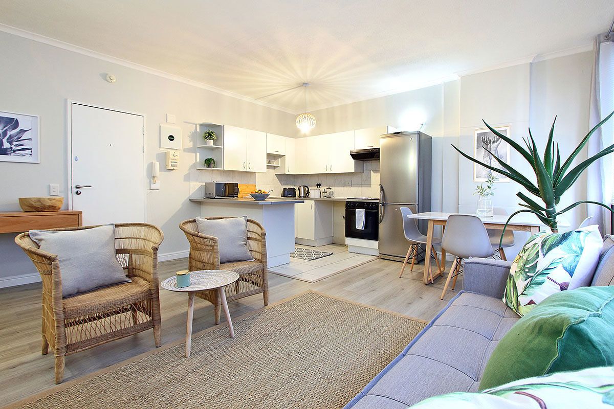 Un salon moderne avec un canapé, deux fauteuils en osier, une petite table ronde et, au fond, une cuisine.