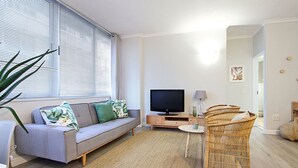 Ein modernes Wohnzimmer mit grauem Sofa, Flachbildfernseher, Korbsessel und großem Fenster mit Jalousien.