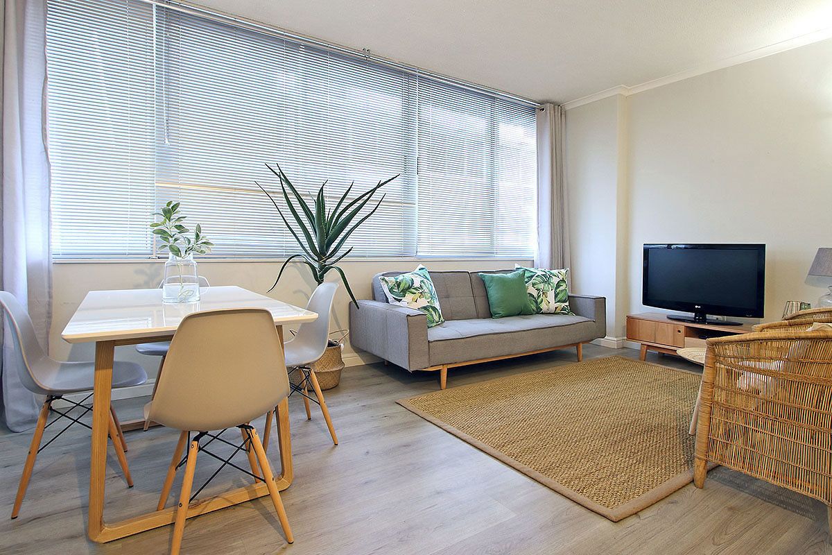 A modern living room with a dining area, a sofa with cushions, a television, and a large window with blinds.