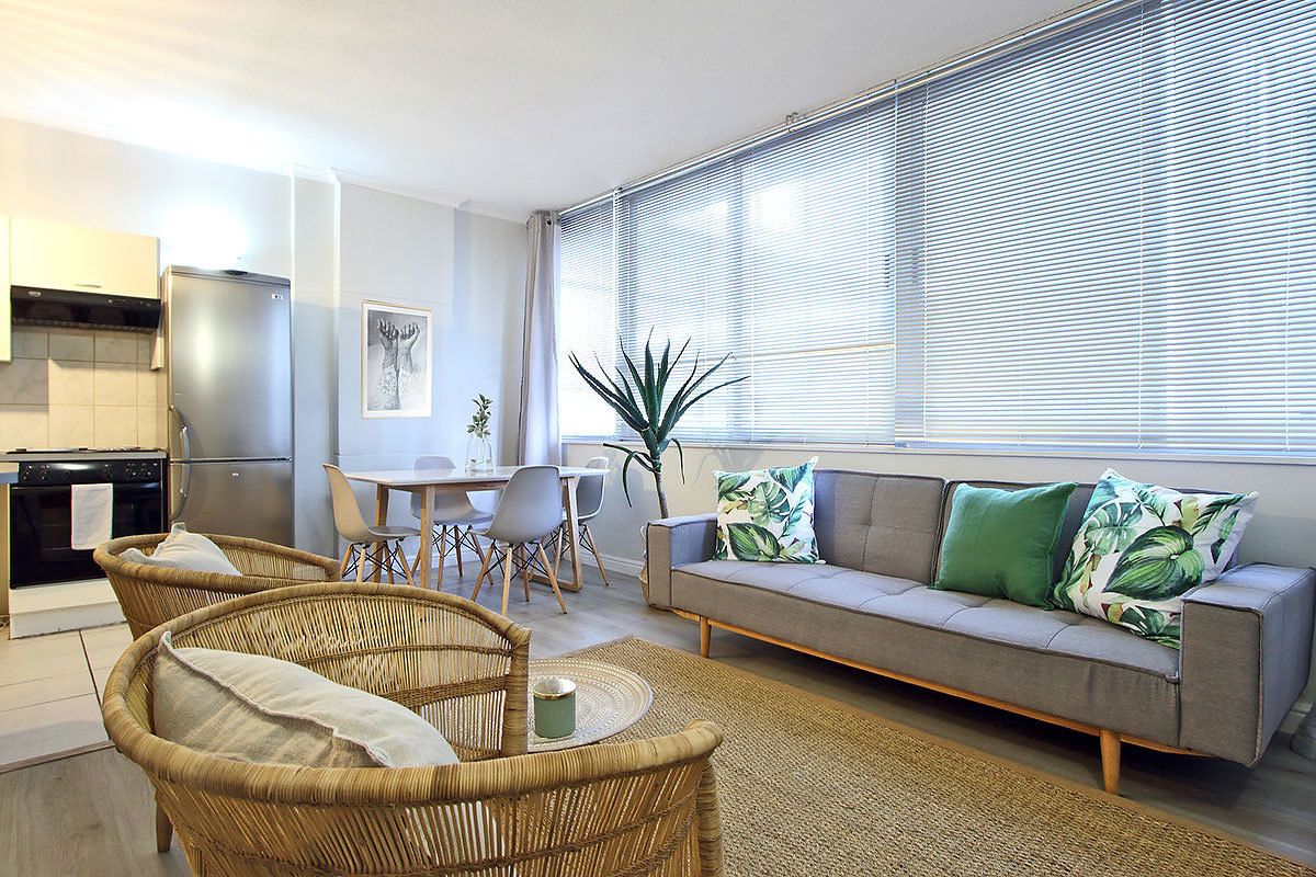 A modern living room with a sofa, two wicker chairs, a dining table with chairs, a kitchen area with a refrigerator, and a large window with blinds.