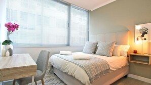 Ein modernes Schlafzimmer mit Bett, Nachttisch, Sessel und einem Fenster mit Jalousien.