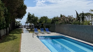 A rectangular swimming pool with a tiled edge, surrounded by a stone wall and a grassy area. There are several blue lounge chairs aligned along the poolside. - Villa Angela (Corfu)