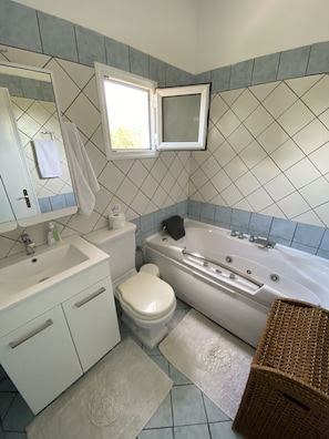Un baño con lavamanos, inodoro y bañera blancos. Hay una ventana sobre la bañera y un espejo sobre el lavamanos.