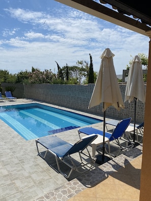 Una piscina con baldosas azules, rodeada de sillas de descanso y sombrillas, con un muro de piedra y follaje al fondo.