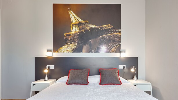 Hotellihuone, jossa on sänky, kaksi yöpöytää, suuri Eiffel-tornin juliste ja punavalkoraidallinen peitto.
