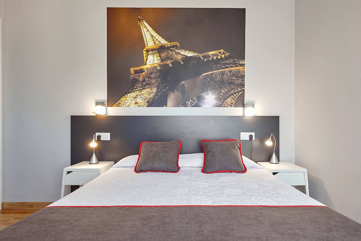 A hotel room with a bed, two nightstands, a large poster of the Eiffel Tower, and a red and white striped blanket.