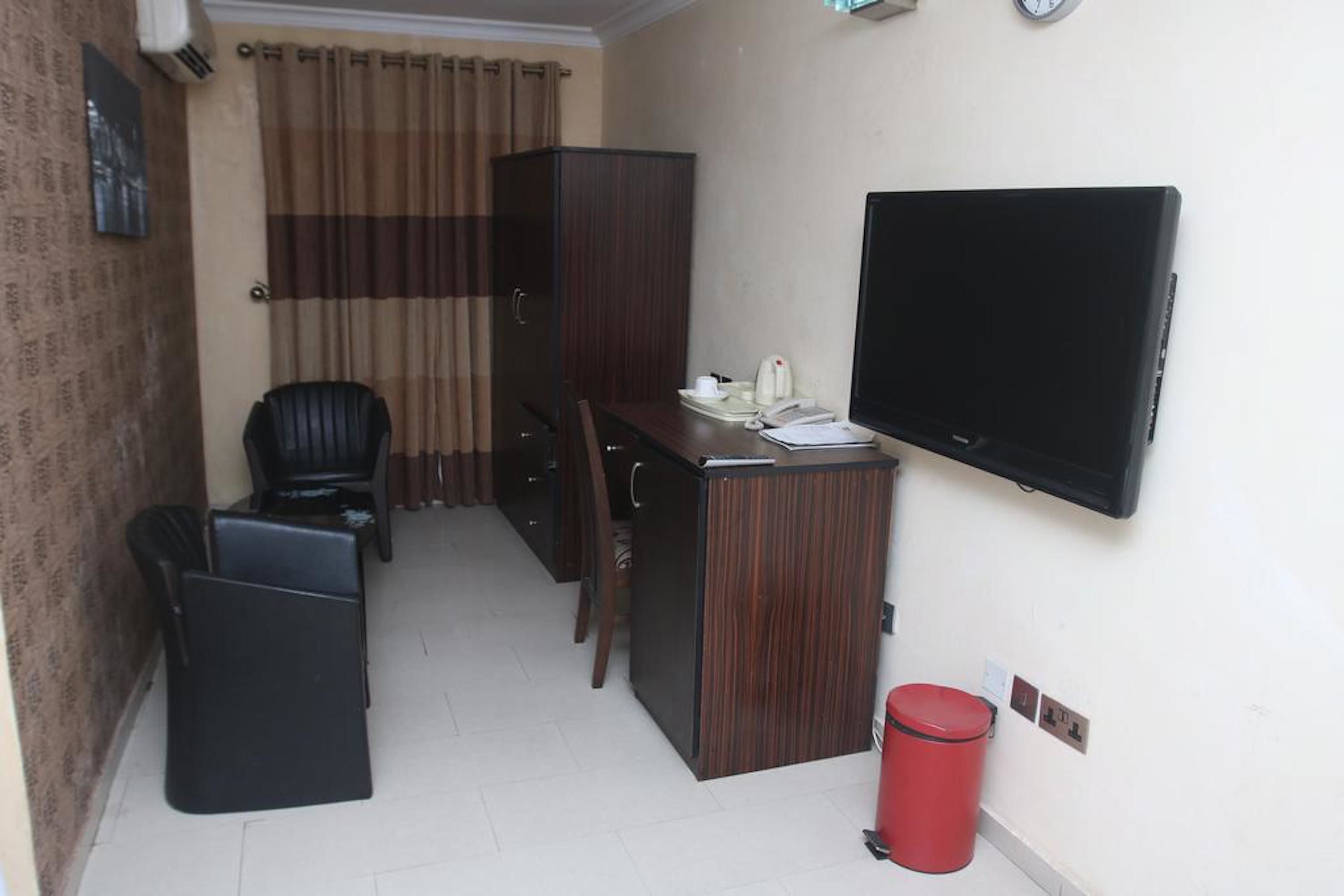 A compact hotel room with a wooden desk, a black chair, a television mounted on the wall, a red trash bin, and a curtain-covered window.
