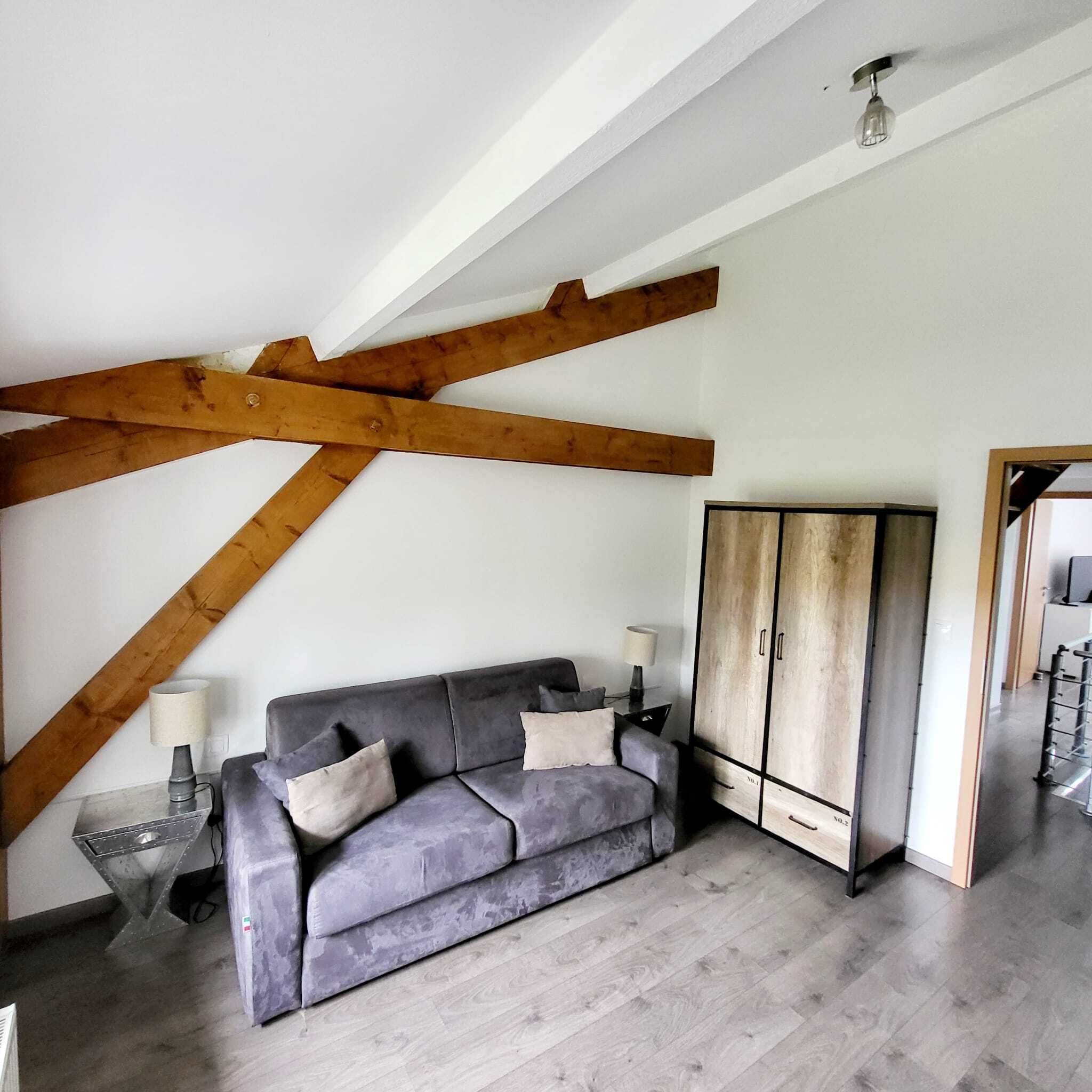 A room with a grey sofa, a wooden ceiling beam, a wardrobe, and a lamp on a side table.