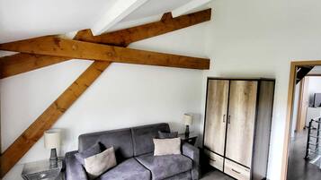 A room with a grey sofa, a wooden ceiling beam, a wardrobe, and a lamp on a side table.
