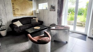 Ein modernes Wohnzimmer mit einer Couch, einem Couchtisch und zwei Sesseln. Ein großes Fenster bietet Blick auf Grünflächen außerhalb.