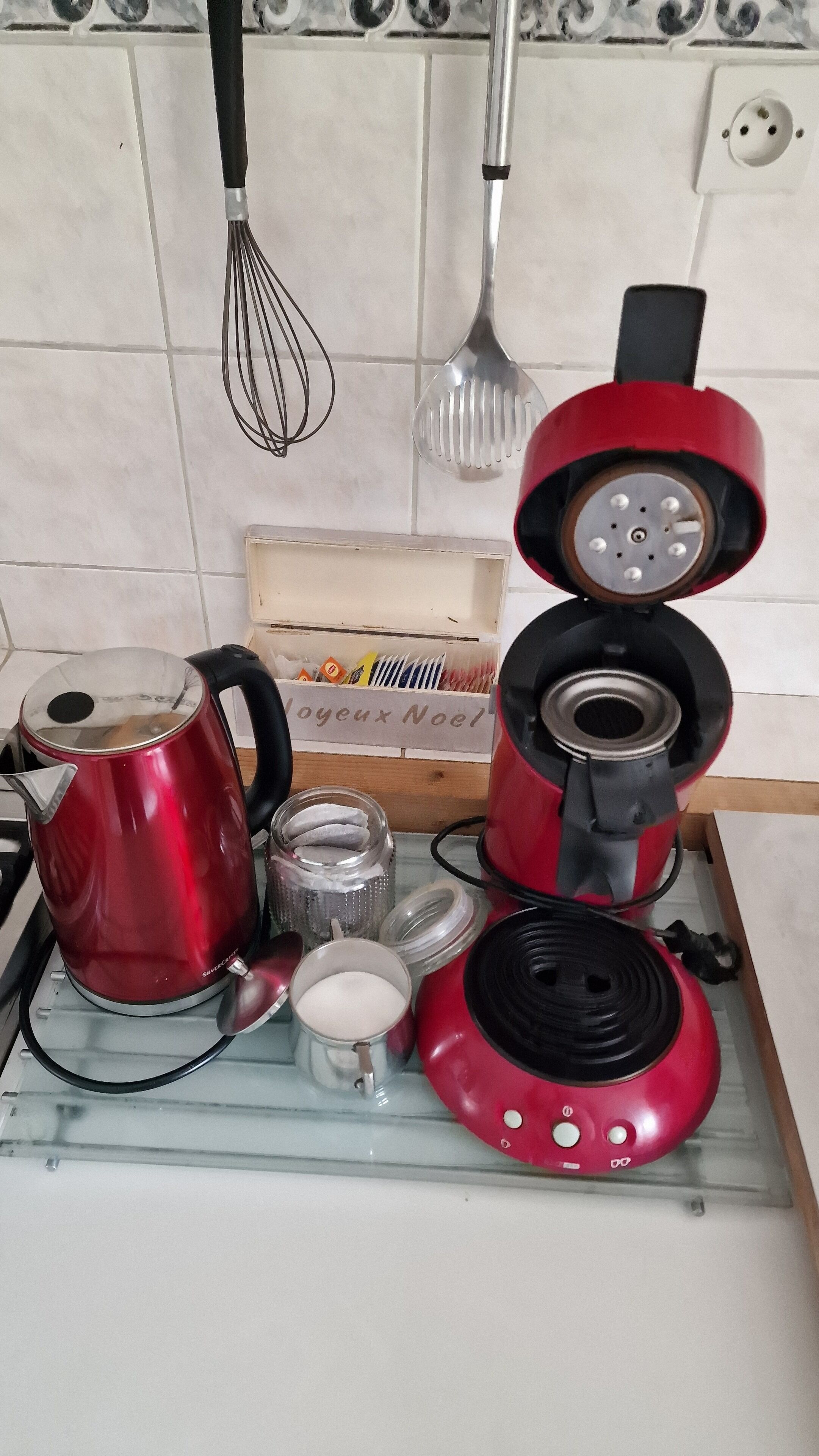 A kitchen counter with a red kettle, a red electric kettle, a glass jar, and a small metal container.