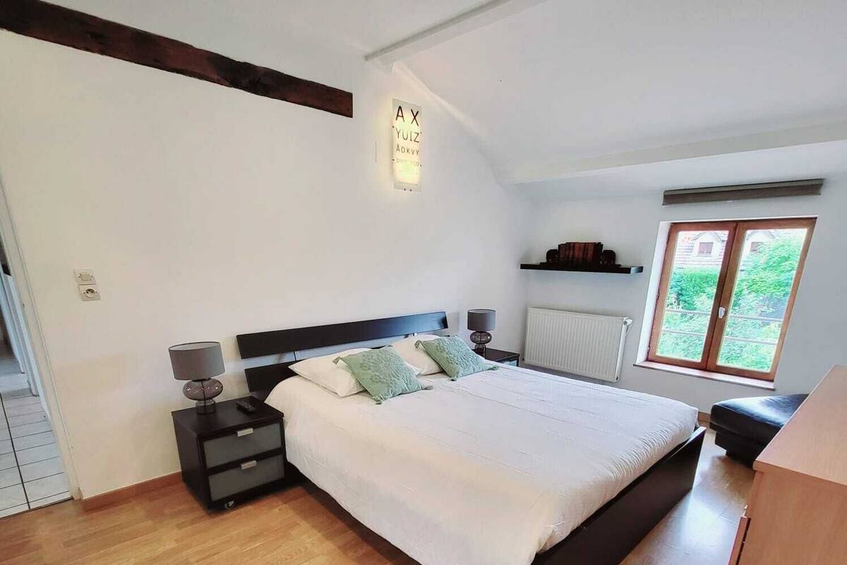 Ein Schlafzimmer mit einem großen Bett, einem Nachttisch, einer Kommode und einem Fenster mit Blick auf Grünflächen.