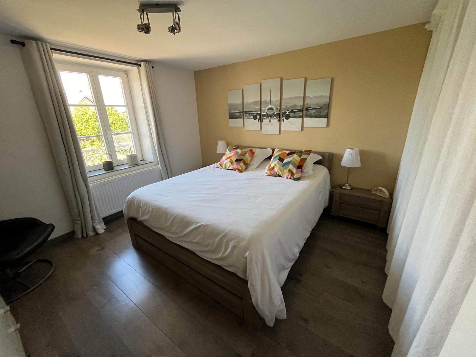 A bedroom with a large bed, a window with curtains, a radiator, a bedside table with a lamp, and a wall with framed artwork.