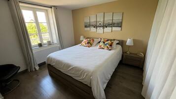 A bedroom with a large bed, a window with curtains, a radiator, a bedside table with a lamp, and a wall with framed artwork.