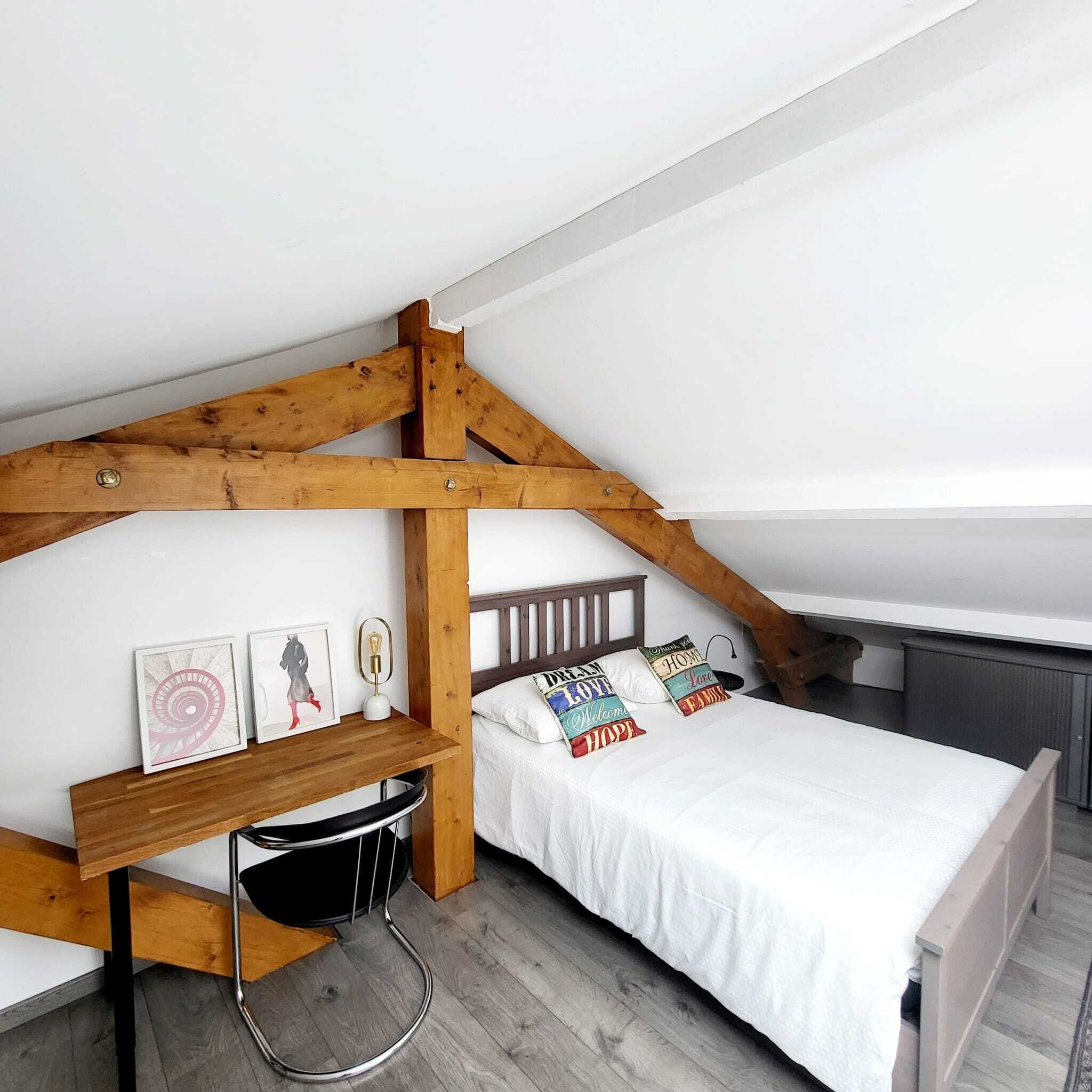 A loft bedroom with a sloped ceiling, wooden beams, a bunk bed, a small desk, and a chair.