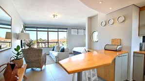 A modern apartment with a kitchen, dining area, and living room. It features large windows, a dining table, a sofa, and a kitchen counter. - Morakani (Cape Town)